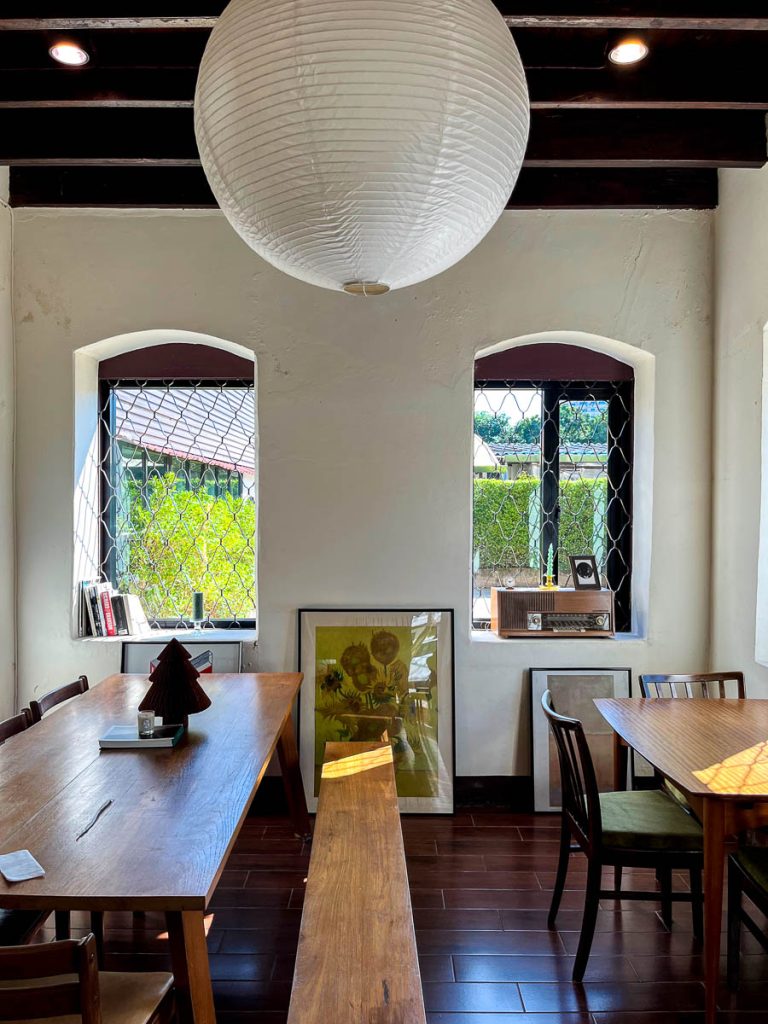 Communal wooden table and bench inside Baan 104, with sunlight streaming through lattice windows and framed artwork leaning against the wall