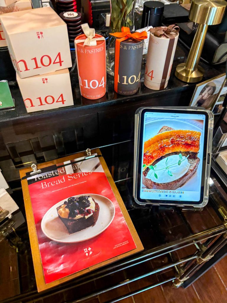 Close-up of The Pantry counter at Baan 104 with branded gift packaging, a tablet displaying banana toast, and a menu board featuring toasted bread options