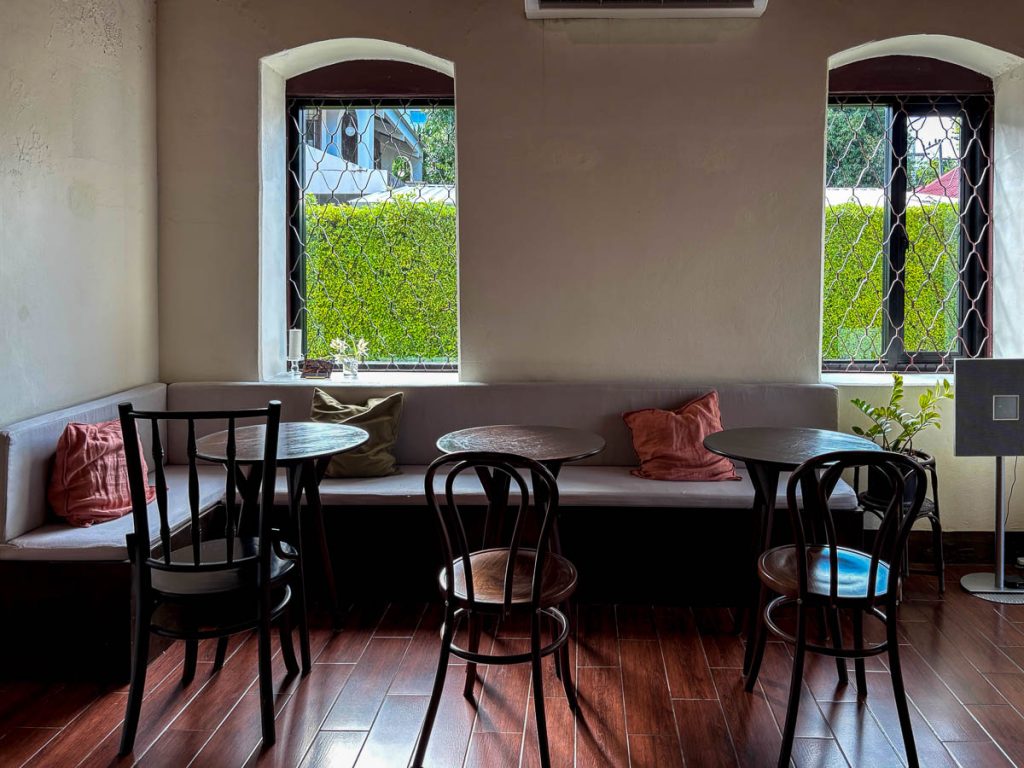 Interior seating at The Pantry inside Baan 104, showing a long grey bench with pillows and round wooden tables positioned beside bright windows with a hedge outside