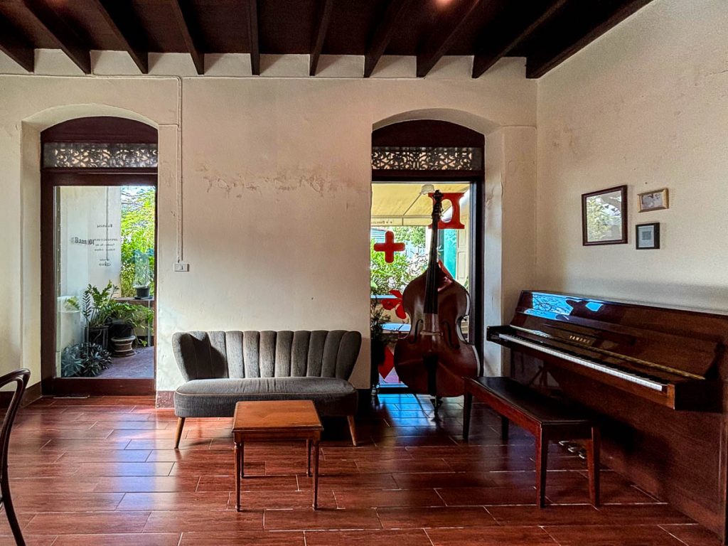 Interior of The Pantry inside Baan 104 showing a grey mid-century sofa, a wooden coffee table, upright piano and double bass, with sunlight coming through doorways and timber flooring